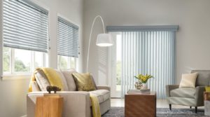 Seafoam blue vertical blinds on a set of patio doors on focal wall with two sets of matching-color window blinds on an adjoining wall in a sitting room with with a light tan sofa, blue-gray easy chair and brown coffee table