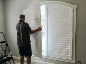 Installer shown from behind putting the second of two custom arched shutters in place over an arch-top window