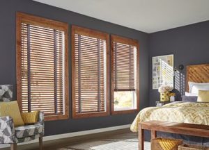 In a bedroom, a set of three large windows, each covered by wood blinds, with one partially raised