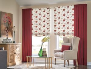 Corner of a sitting area with an off-white Queen Anne wingback chair holding a red pillow in front of windows covered by red-and-white floral pattern Roman shades that are flanked by solid-red, floor-length drapes