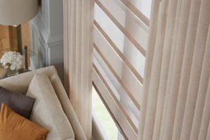 Tan-and-white roller shades with sheer effects layered with matching tan pleated drapes