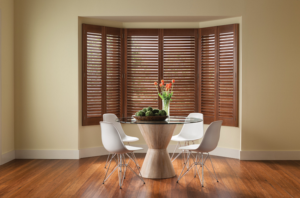 Finished wood shutters with louvers closed on a bay window behind a glass-top table and chairs