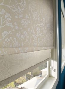 Layered roller shades on a window, with a beige and white tree-patterned shade over a tan, open-weave shade